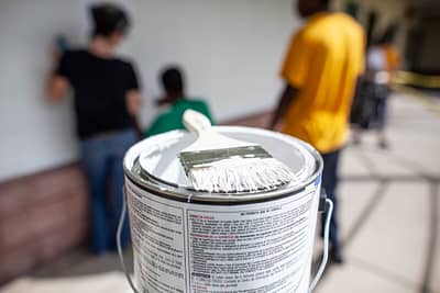 Closeup of a paintbrush on a paint can with volunteers working in the background.