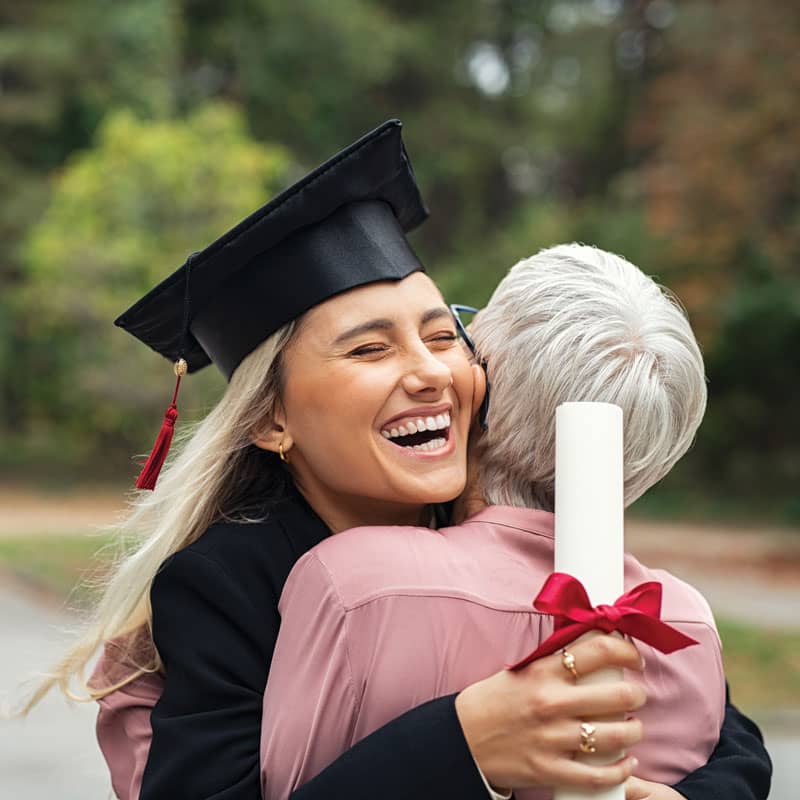 A young woman in her cap and gown smiles and hugs her grandmother with her diploma in hand.