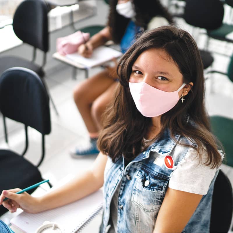 A teenage girl wearing a pink mask sits at a desk in a classroom.
